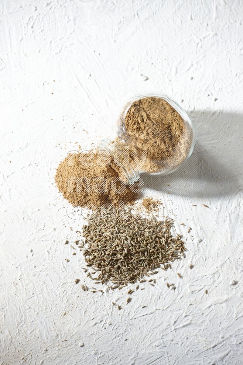 A flipped glass spice jar full of cumin powder and powder spilled out with cumin seeds on textured white flooring