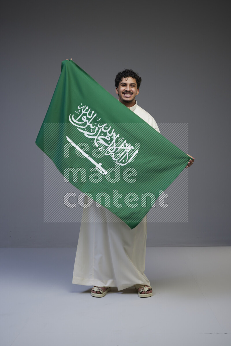 A Saudi man standing wearing thob holding big Saudi flag on gray background