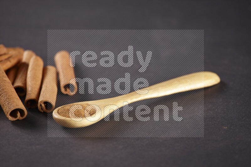 Cinnamon sticks stacked beside a wooden spoon full of cinnamon powder on black background