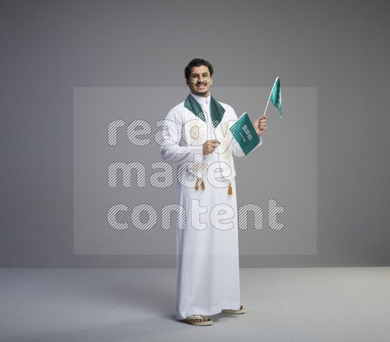 A Saudi man standing wearing thob and saudi flag scarf with face painting holding small Saudi flag on gray background