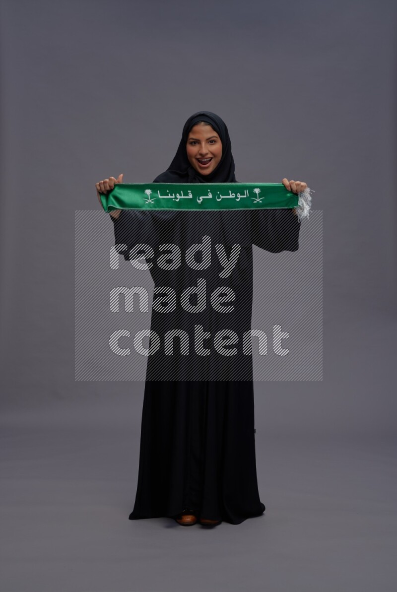 Saudi woman wearing Abaya standing holding Saudi flag on gray background