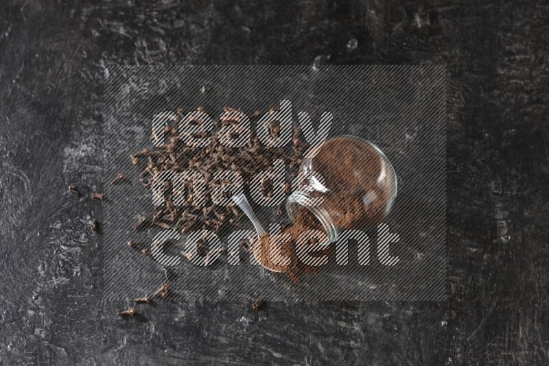 A flipped glass spice jar and a metal spoon full of cloves powder with cloves spread on textured black flooring