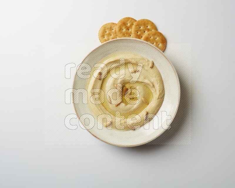 Hummus in a pottry plate garnished with pine nuts on a white background