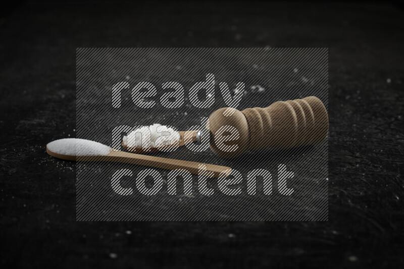 2 wooden spoons filled with fine and coarse salt with wooden grinder beside them on black background