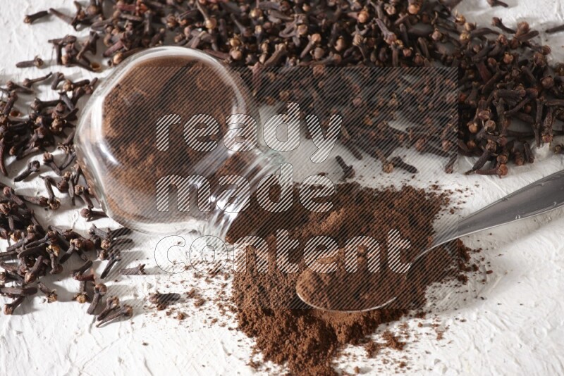 A flipped glass spice jar and a metal spoon full of cloves powder and powder came out of the jar with cloves spread on textured white flooring