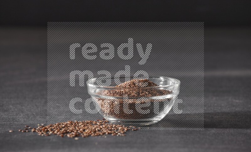 A glass bowl full of flaxseeds surrounded by the seeds on a black flooring