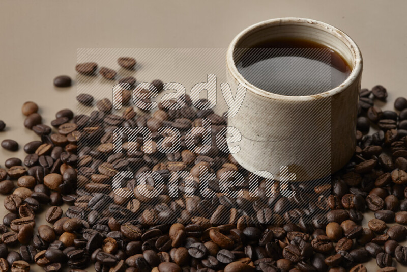 A beige pottery cup of coffee surrounded by roasted coffee beans on beige background
