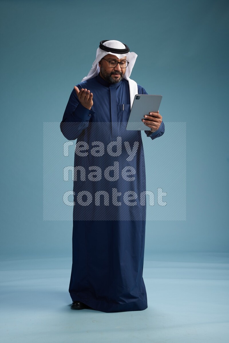 Saudi Man with shimag Standing holding ATM card while working on tablet on blue background