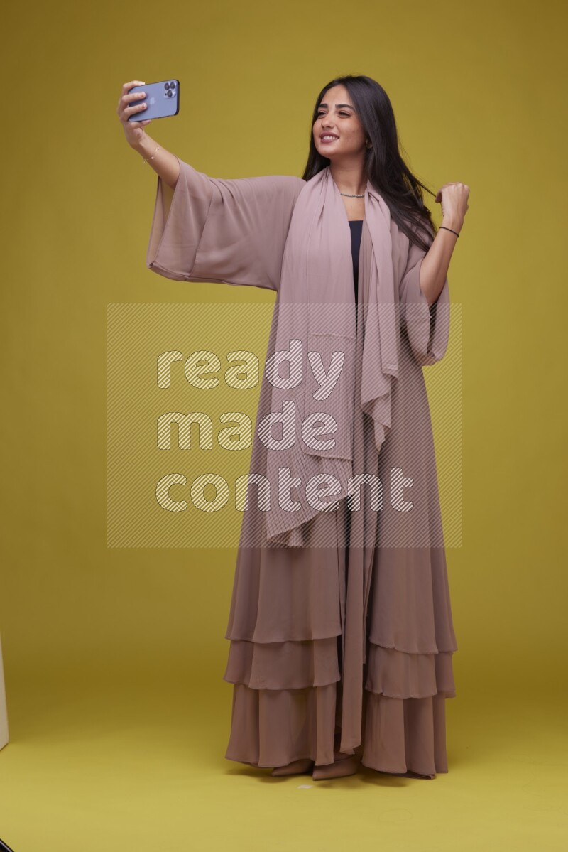 A woman Taking a Selfie on a Yellow Background wearing Brown Abaya