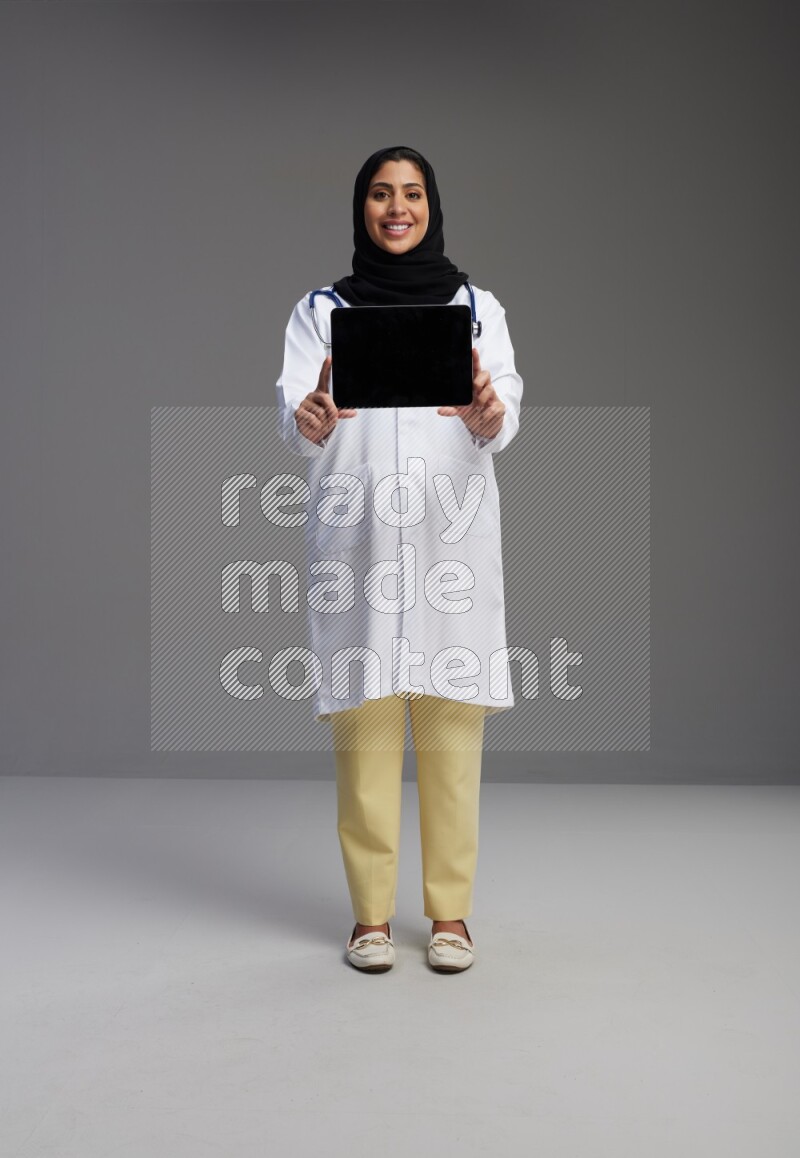 Saudi woman wearing lab coat with stethoscope standing showing tablet to camera with sign in the back on Gray background