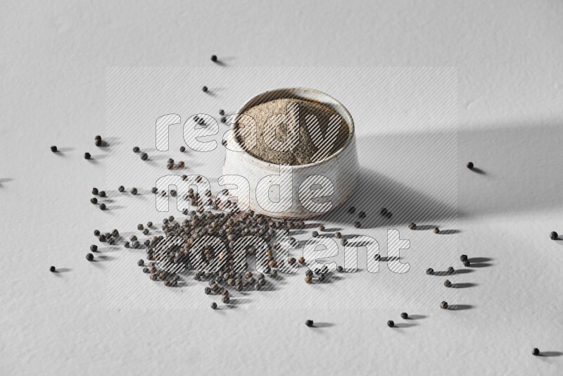 A white ceramic bowl full of black pepper powder and black pepper beads spread on white flooring