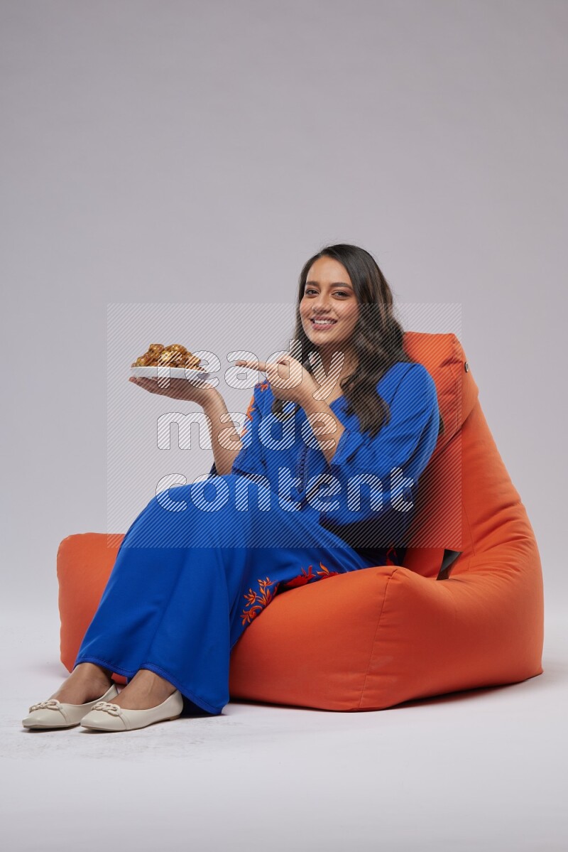 A Woman sitting on an orange beanbag wearing Jalabeya holding a plate of dates