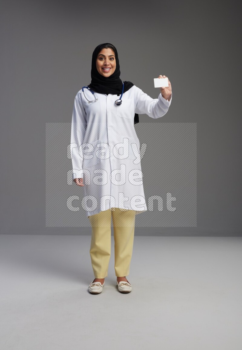 Saudi woman wearing lab coat with stethoscope standing holding ATM card on Gray background