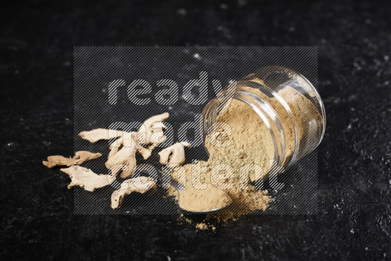 A glass jar full of ground ginger powder flipped with some spilling powder on black background