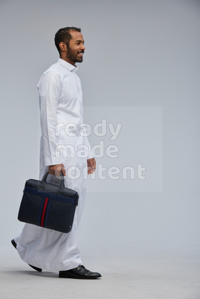 Saudi man wearing thob standing holding suitcase on gray background