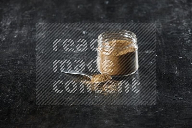 A glass jar and a metal spoon full of cumin powder on a textured black flooring