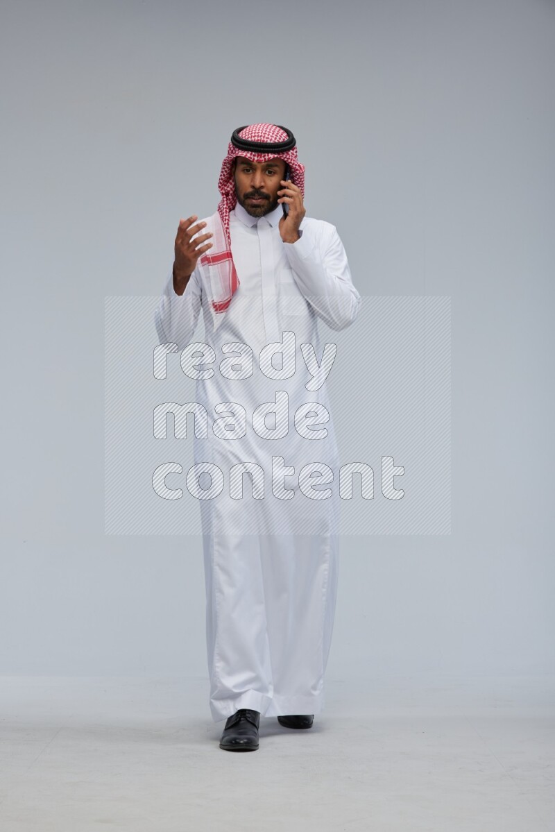 Saudi man Wearing Thob and shomag standing talking on phone on Gray background