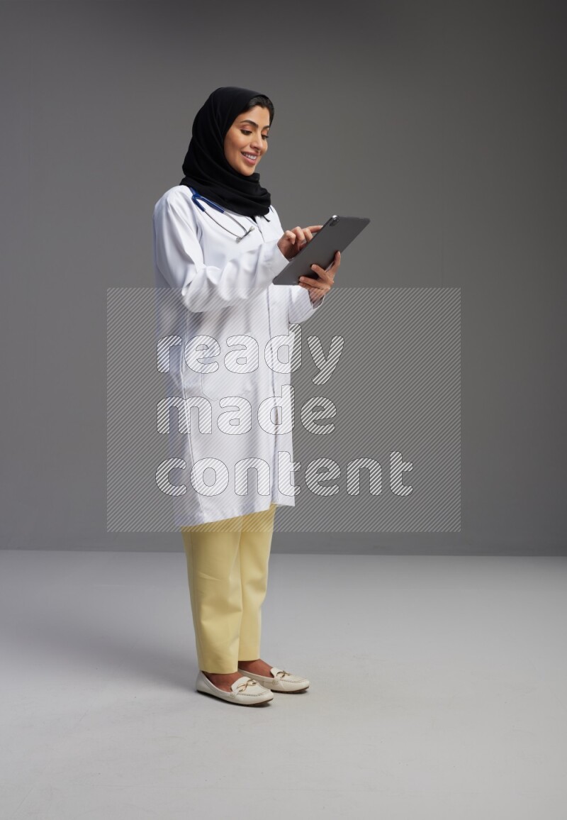Saudi woman wearing lab coat with stethoscope standing working on tablet on Gray background