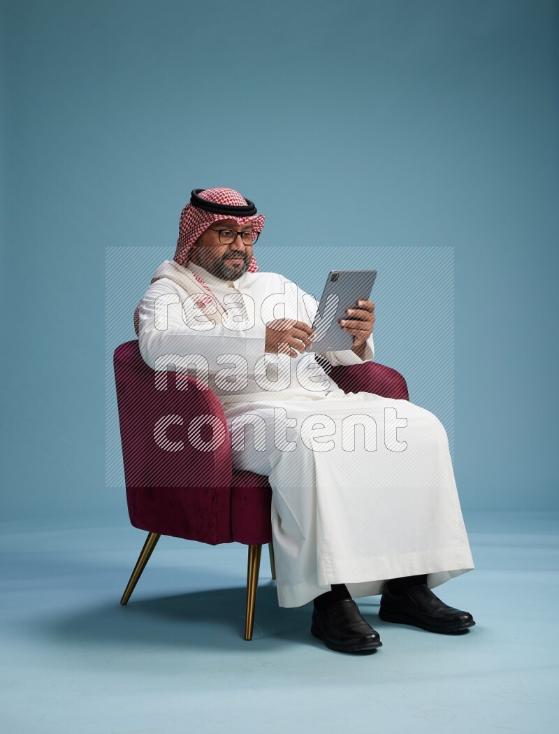 Saudi Man with shimag sitting on chair working on tablet on blue background