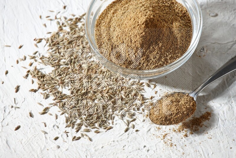 وعاء زجاجي وملعقة معدنية ممتلئان ببودرة الكمون وحبوب الكمون تحتهما على خلفية بيضاء