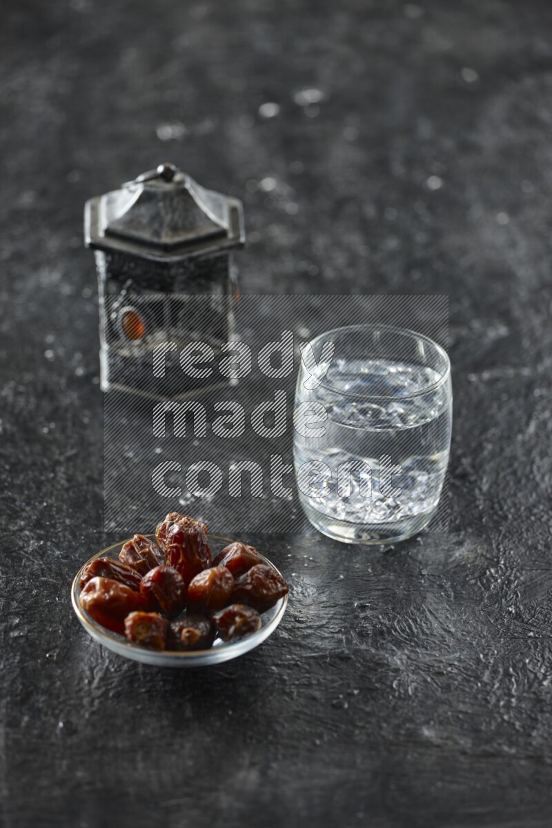 A silver lantern with different drinks, dates, nuts, prayer beads and quran on textured black background