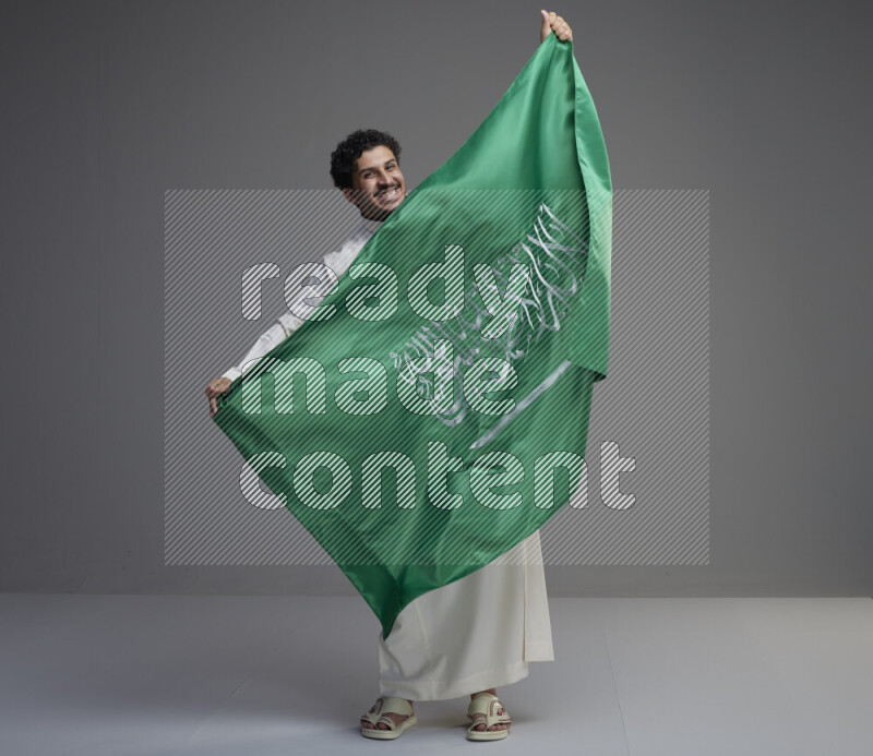 A Saudi man standing wearing thob holding big Saudi flag on gray background