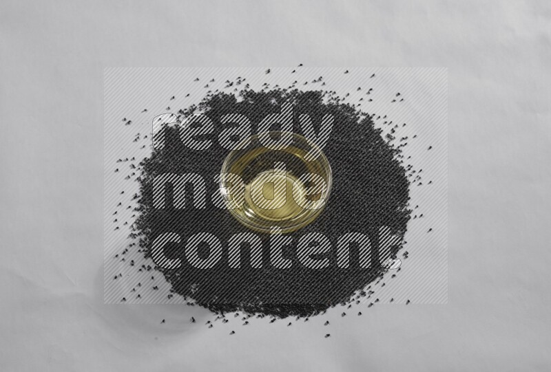 A glass bowl full of black seeds oil surrounded by the seeds on a white flooring