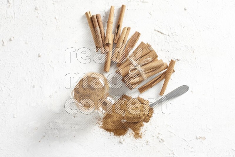 Flipped herbs glass jar full of cinnamon powder with a metal spoon full of powder and cinnamon sticks on a textured white background