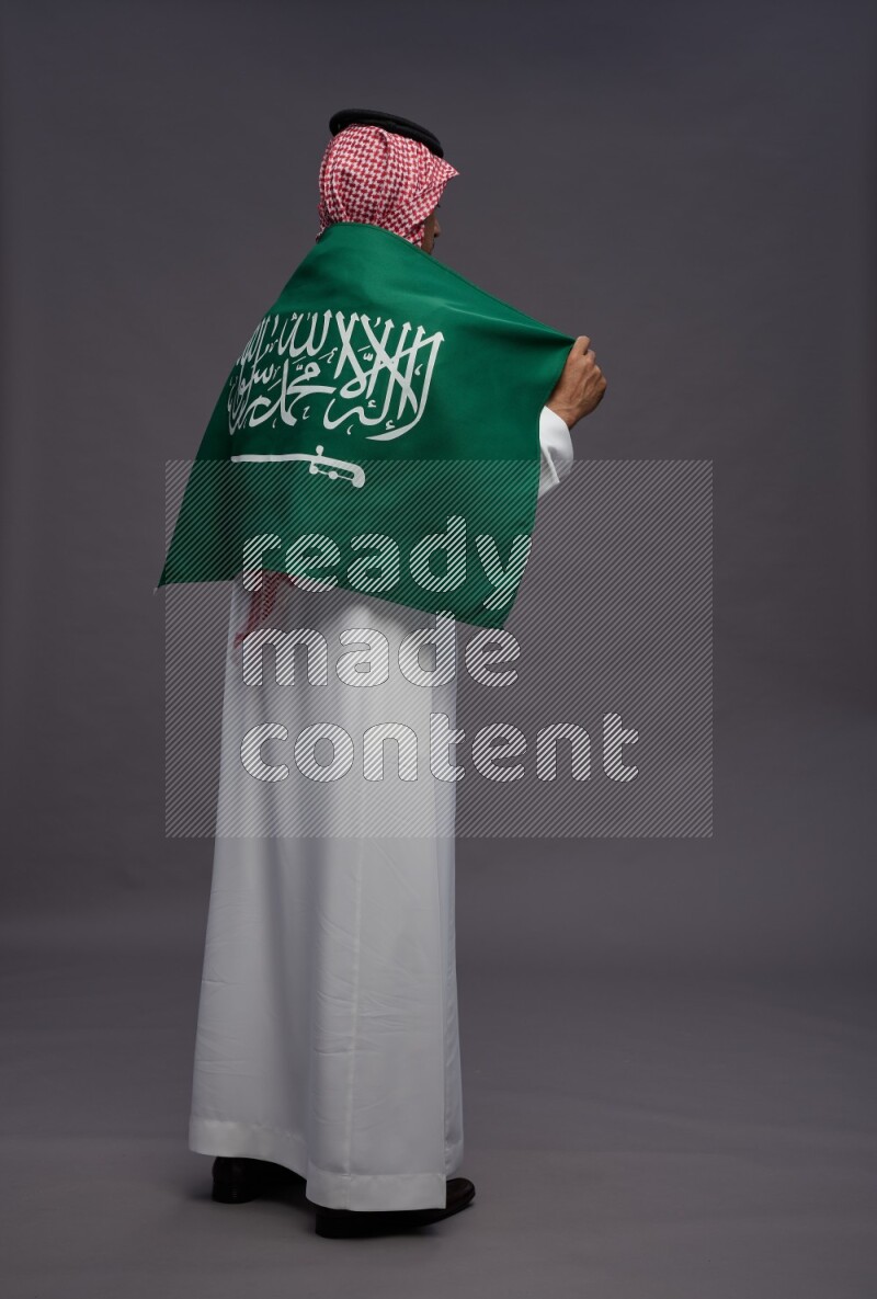 Saudi man wearing thob and shomag standing holding Saudi flag on gray background