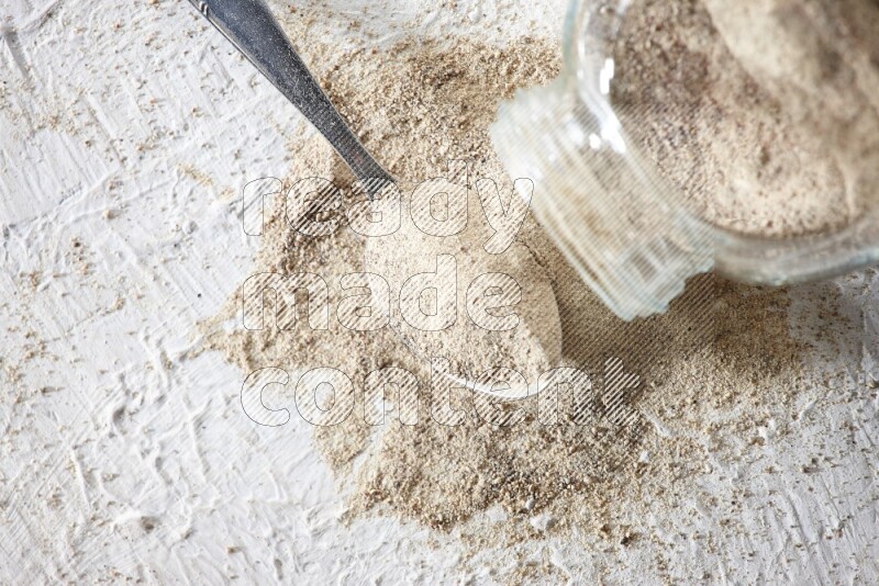 A flipped herbal glass jar and metal spoon full of white pepper powder with spilled powder on textured white flooring
