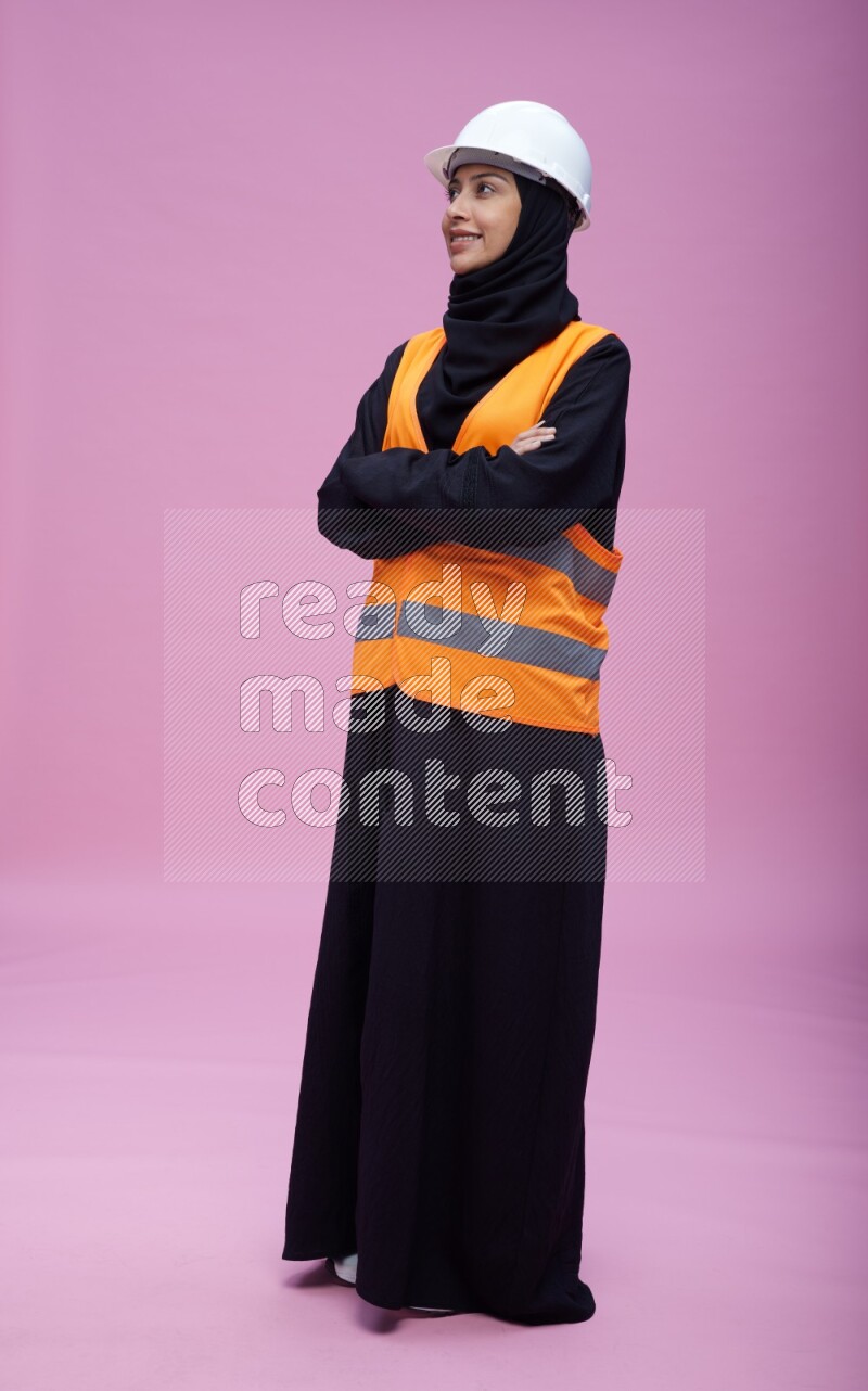 Saudi woman wearing Abaya with engineer vest and helmet standing with crossed arms on pink background