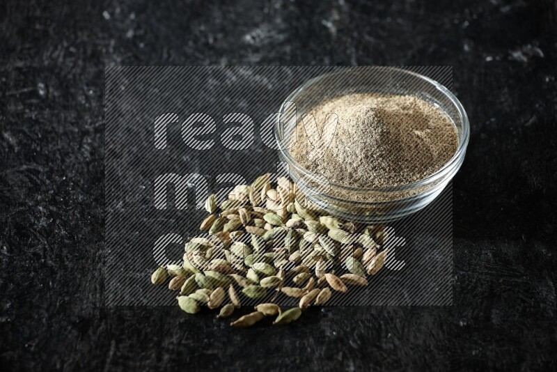 A glass bowl full of cardamom powder and cardamom seeds beside it on textured black flooring
