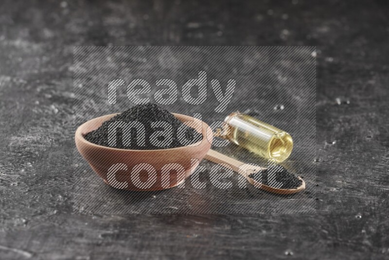 A wooden bowl and spoon full of black seeds and a bottle of black seeds oil on a textured black flooring