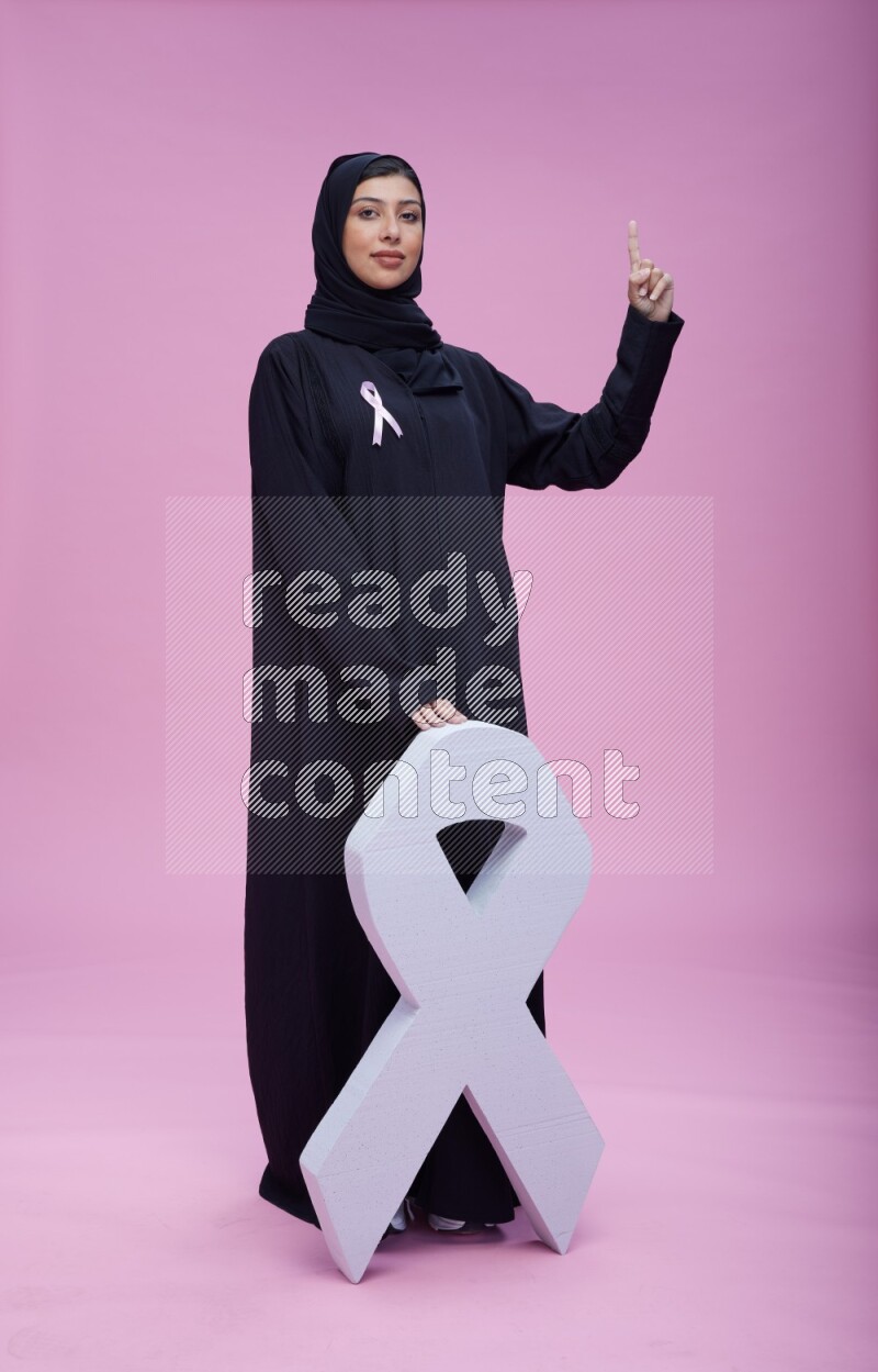 Saudi woman wearing pink ribbon on abaya standing holding awareness ribbon on plain pink background