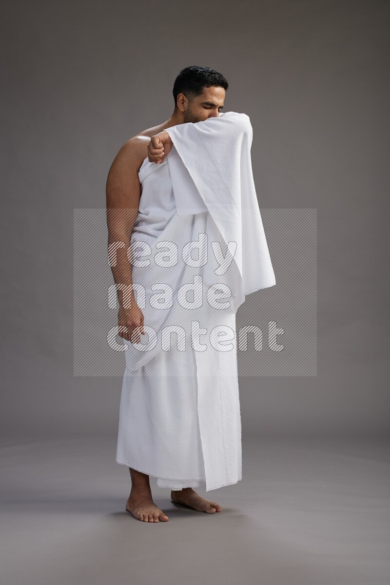 A man wearing Ehram standing showing how to sneeze on gray background