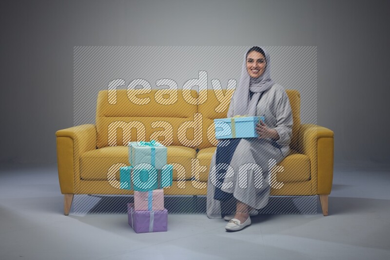 A Saudi woman wearing a light gray Abaya and head scarf sitting on a yellow sofa holding a gift box beside multi color gift boxes eye level on a grey background