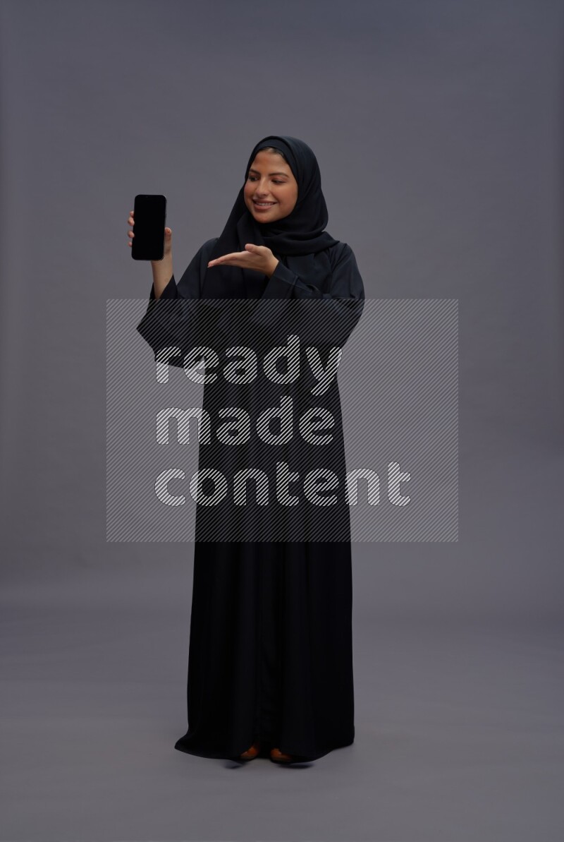 Saudi woman wearing Abaya standing showing phone to camera on gray background