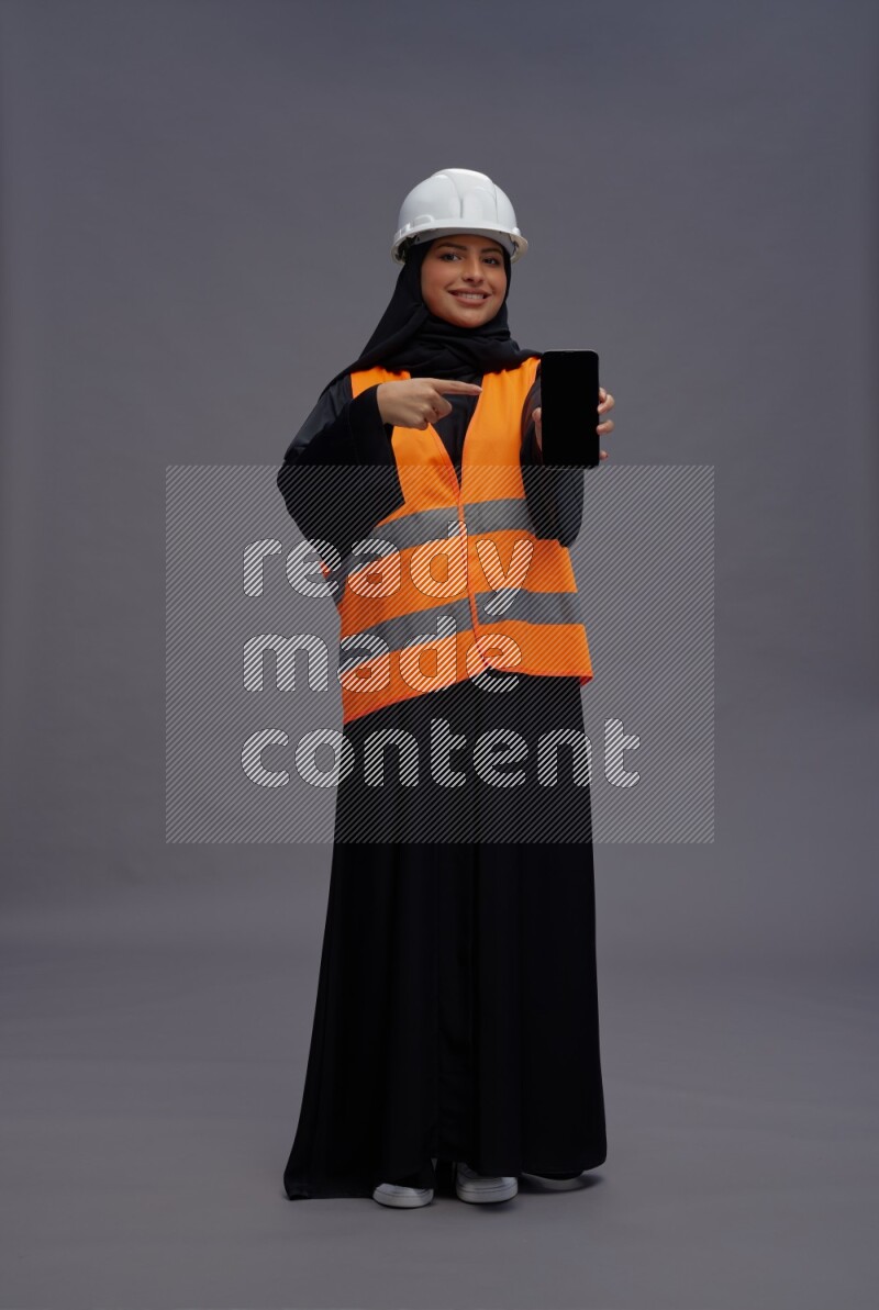 Saudi woman wearing Abaya with engineer vest standing showing phone to camera on gray background