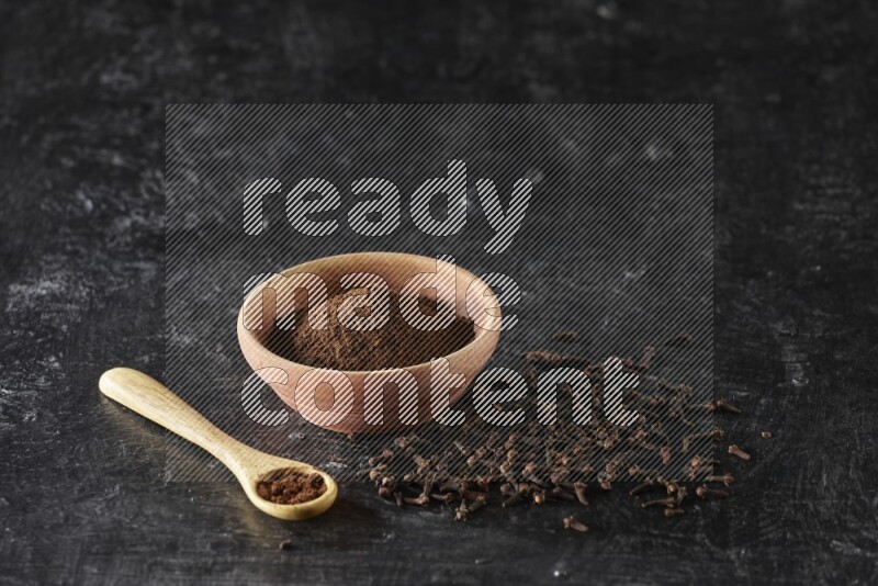 A wooden bowl and wooden spoon full of cloves powder with spreaded cloves on textured black flooring