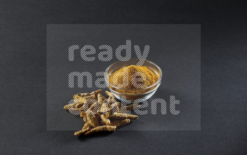 A glass bowl full of turmeric powder and dried whole fingers beside it on black flooring