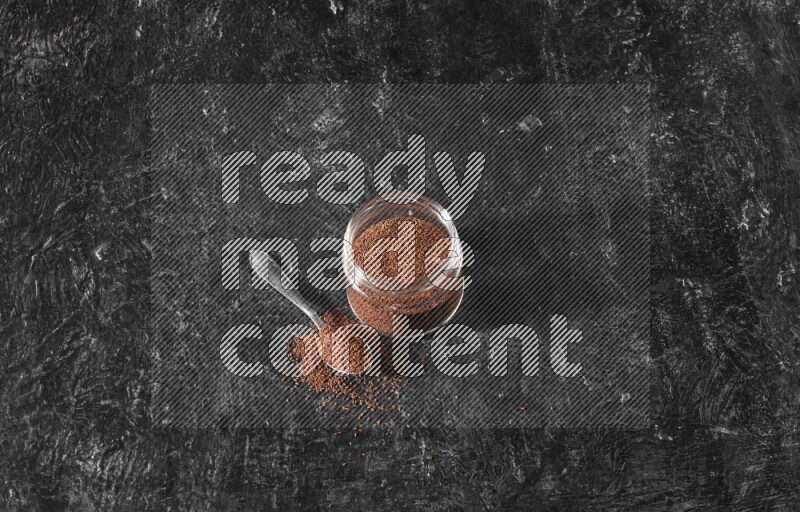 وعاء زجاجي ممتلئ ببذور حب الرشاد مع ملعقة معدنية ممتلئة بالبذور على أرضية سوداء