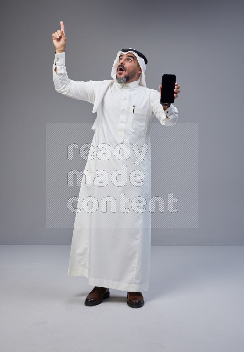 Saudi man Wearing Thob and white Shomag standing showing phone to camera on Gray background
