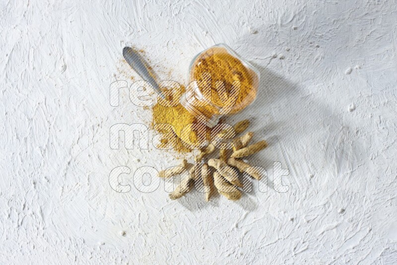 A flipped glass spice jar and metal spoon full of turmeric powder and powder spilled out of it with dried whole fingers on textured white flooring