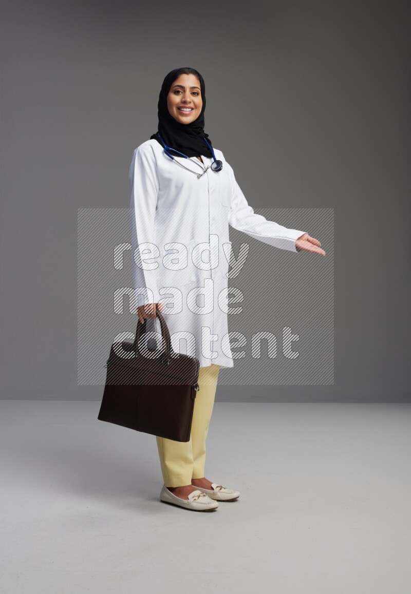 Saudi woman wearing lab coat with stethoscope standing holding bag on Gray background