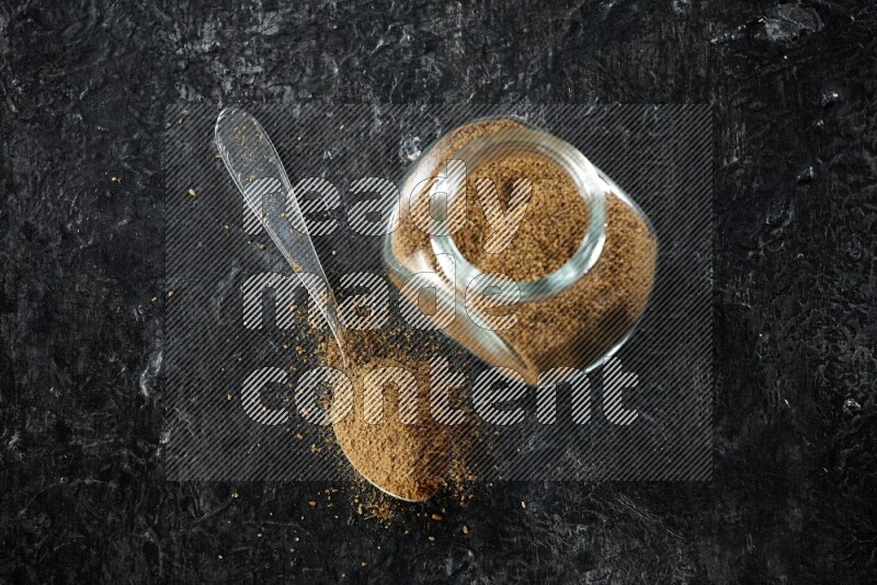 A glass spice jar and a metal spoon full of cumin powder on a textured black flooring