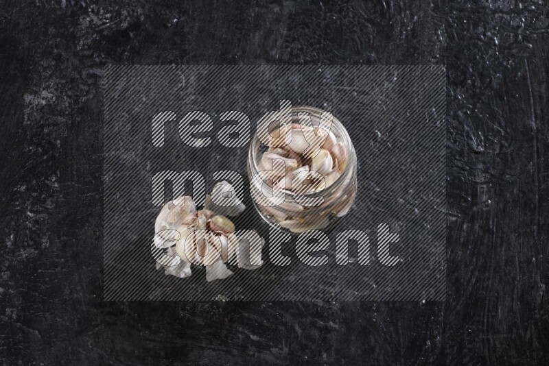 A glass jar full of garlic cloves with a whole garlic bulb beside it on a textured black flooring