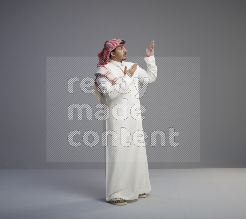 A Saudi man standing wearing thob and red shomag interacting with the camera on gray background