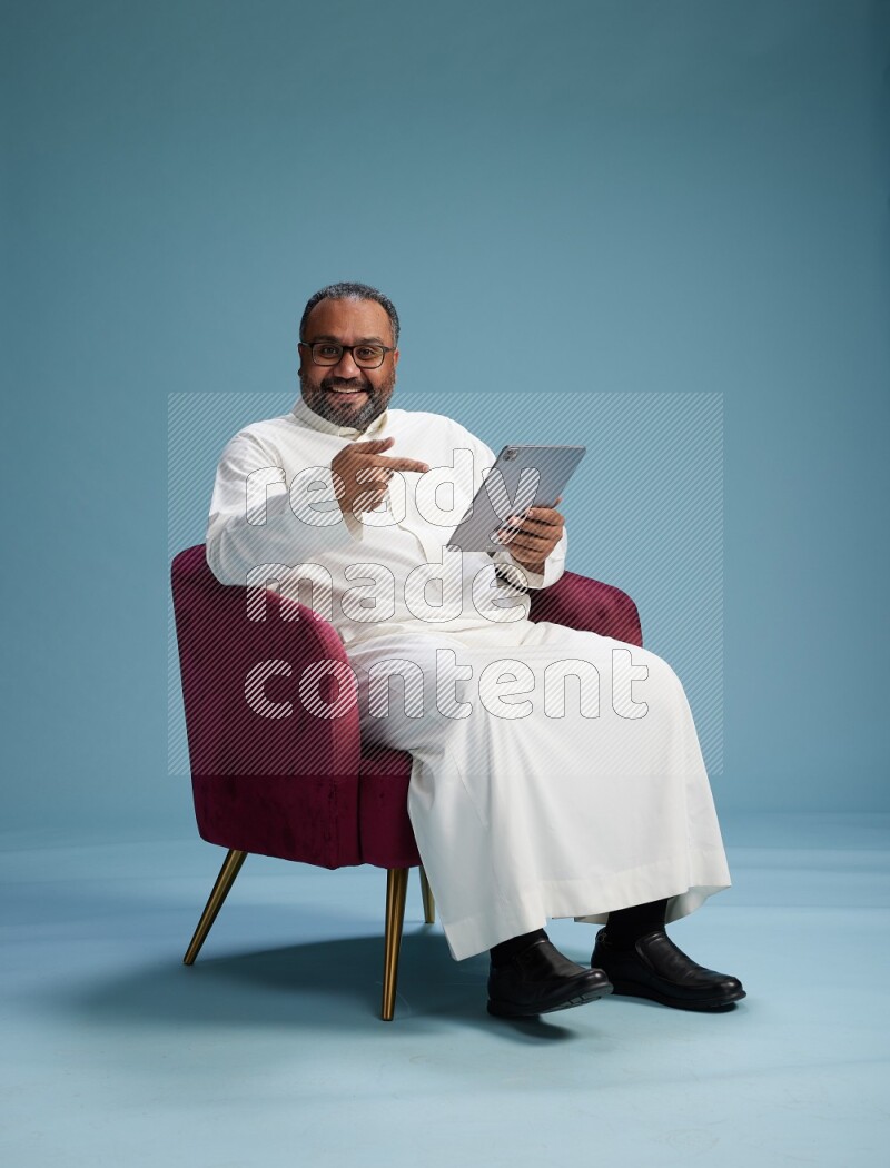 Saudi Man without shimag sitting on chair working on tablet on blue background