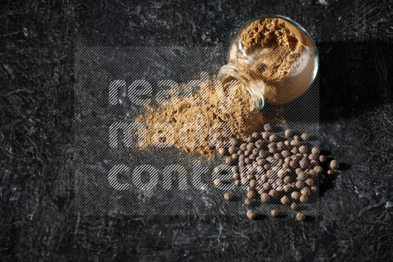 A flipped glass spice jar full of allspice powder and powder spilled out of it with whole balls on a textured black flooring