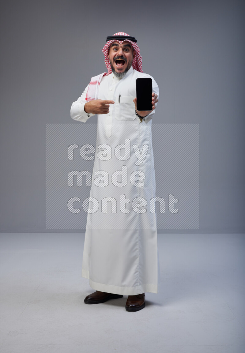 Saudi man Wearing Thob and red Shomag standing showing phone to camera on Gray background